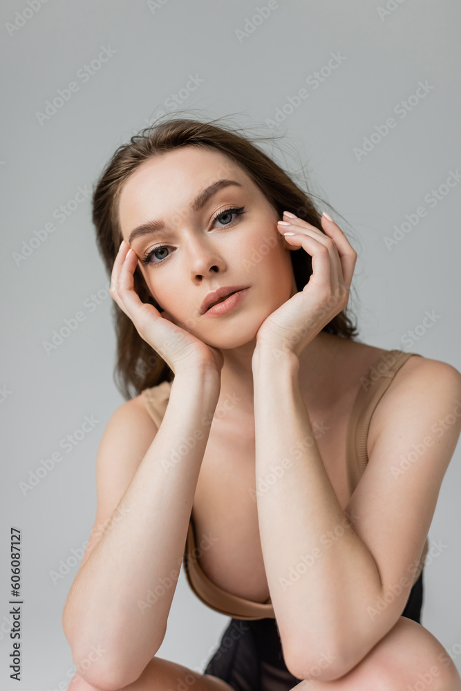 Portrait of young and seductive woman with brunette hair and natural makeup wearing bodysuit while touching face and looking at camera isolated on grey Portrait of young and seductive woman with brunette hair and natural makeup wearing bodysuit while touching face and looking at camera isolated on grey
