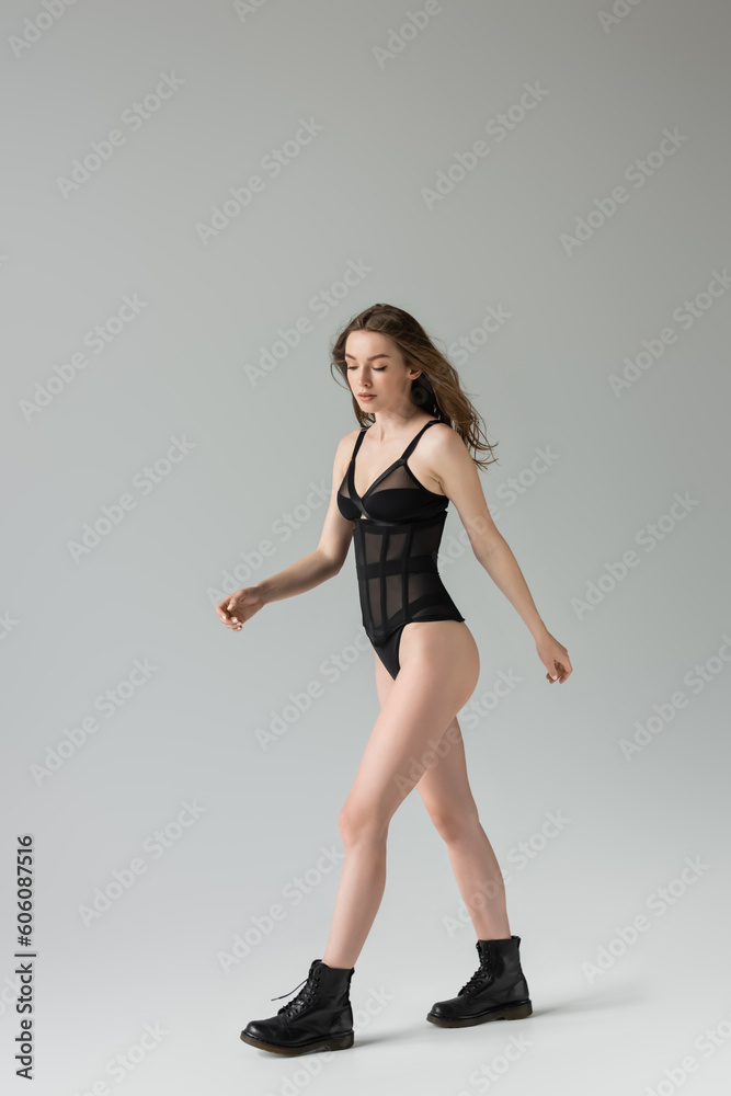 Full length of sensual and fashionable young brunette model with slim body in black bodysuit and stylish boots looking down while walking on grey background
