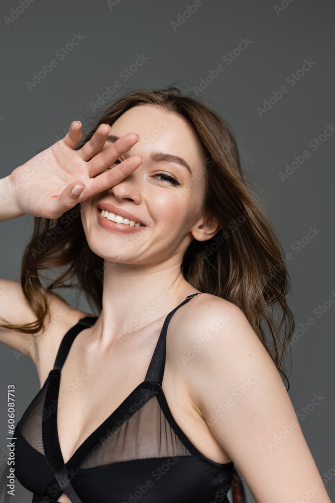 Portrait of young and pretty woman with hairstyle and everyday makeup wearing stylish black bodysuit, smiling and camera and holding hand near face isolated on grey