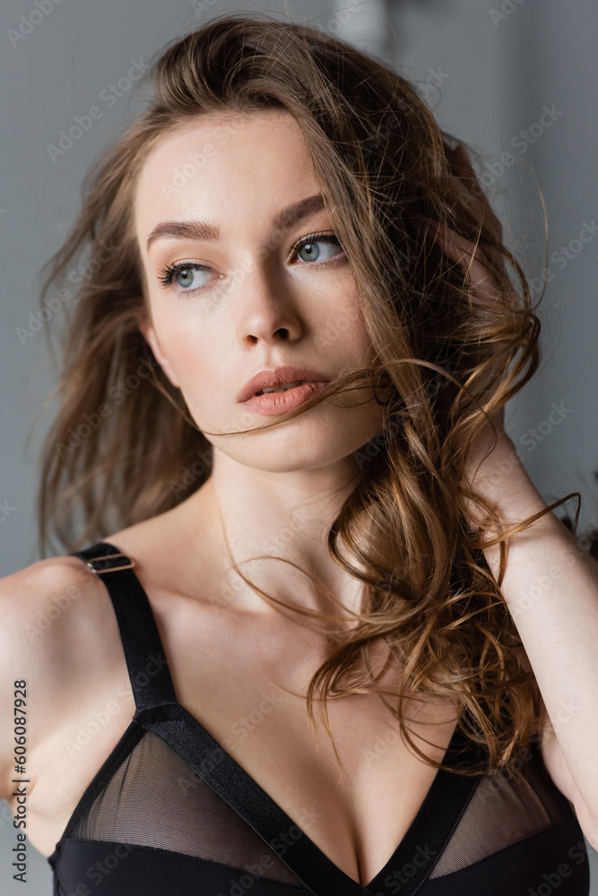 Portrait of young and confident model with brunette hair and natural makeup wearing black bodysuit and looking away while standing on grey background Portrait of young and confident model with brunette hair and natural makeup wearing black bodysuit and looking away while standing on grey background