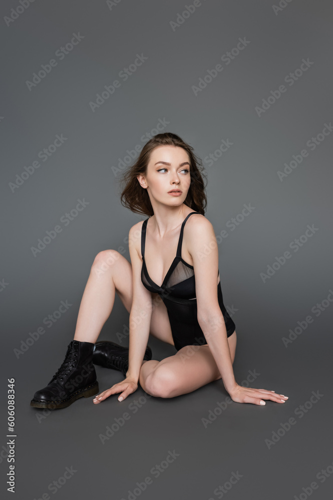 Full length of sexy and slim young brunette woman with natural makeup wearing boots and black bodysuit and looking away while sitting on grey background