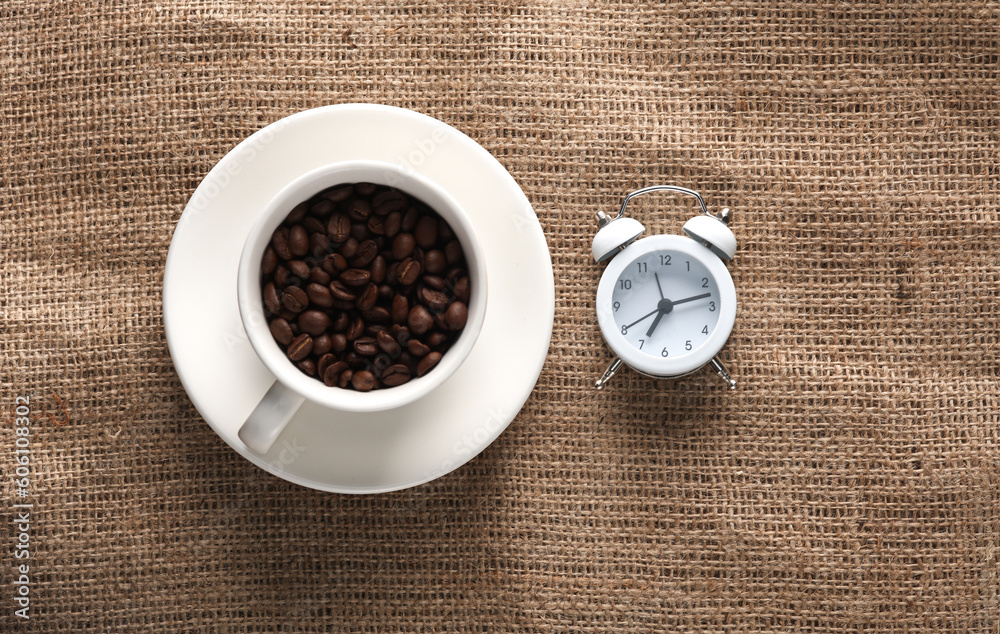 White ceramic cup with coffee beans and alarm clock on burlap. Top view White ceramic cup with coffee beans and alarm clock on burlap. Top view
