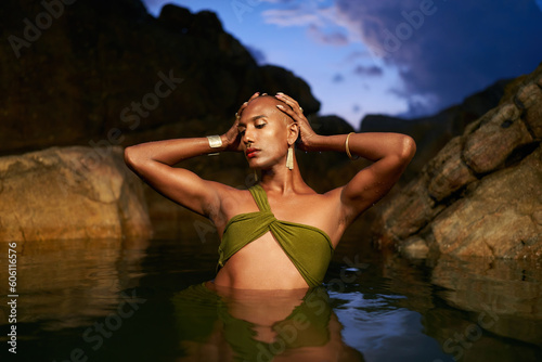 Gender fluid black person dives out from water in natural still water pool. Queer ethnic fashion model in long revealing clothes poses gracefully waist deep in middle of crystal clear lake at night.