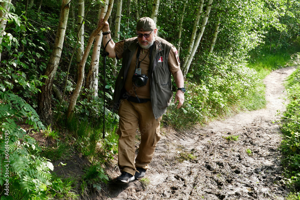 Fototapeta premium Active senior man of 70 with camera and walking stick tries to keep his shoes out of the mud and grabs a tree trunk