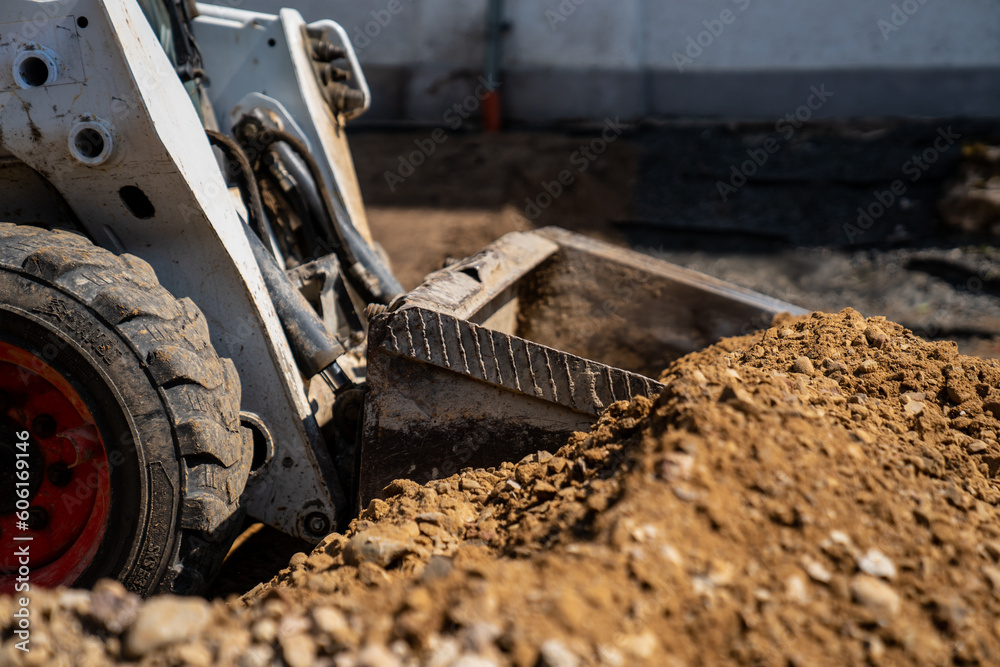 Skid steer loader loading ground and gravel with metal bucket in construction yard next to a building close up