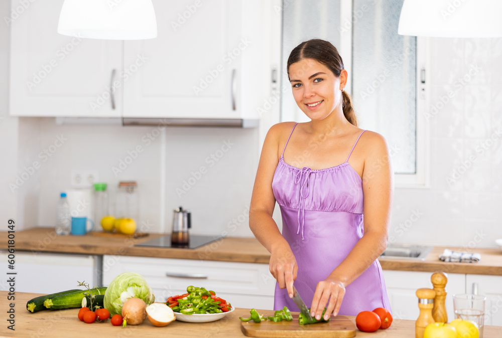 Portrait of positive girl in nightdress standing at kitchen table at home, slicing vegetables, cooking salad.