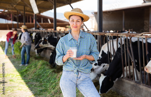 Confident asian woman dairy farm owner standing with glass of fresh milk near stalls with cows on sunny summer day