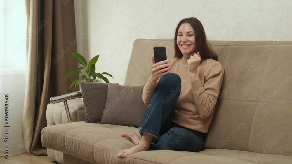 A pretty young adult Caucasian woman uses the phone while sitting on the couch and suddenly solemnly rejoices A pretty young adult Caucasian woman uses the phone while sitting on the couch and suddenly solemnly rejoices