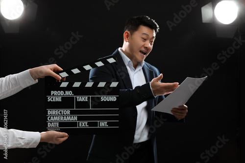 Emotional asian actor performing role while second assistant camera holding clapperboard on stage, selective focus. Film industry