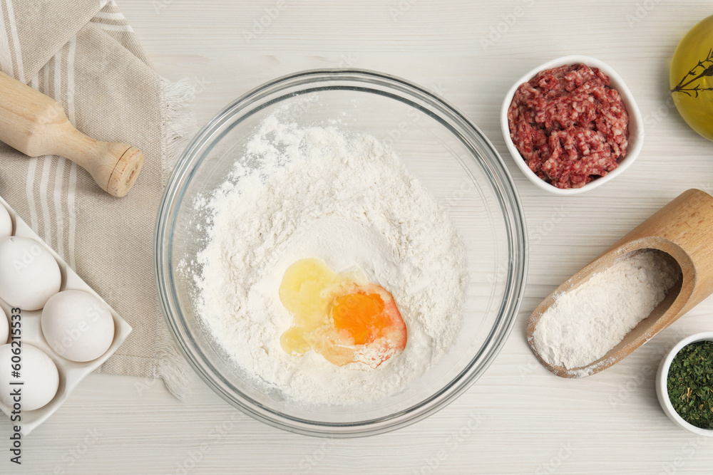 Bowl with flour, egg and products on white wooden table, flat lay