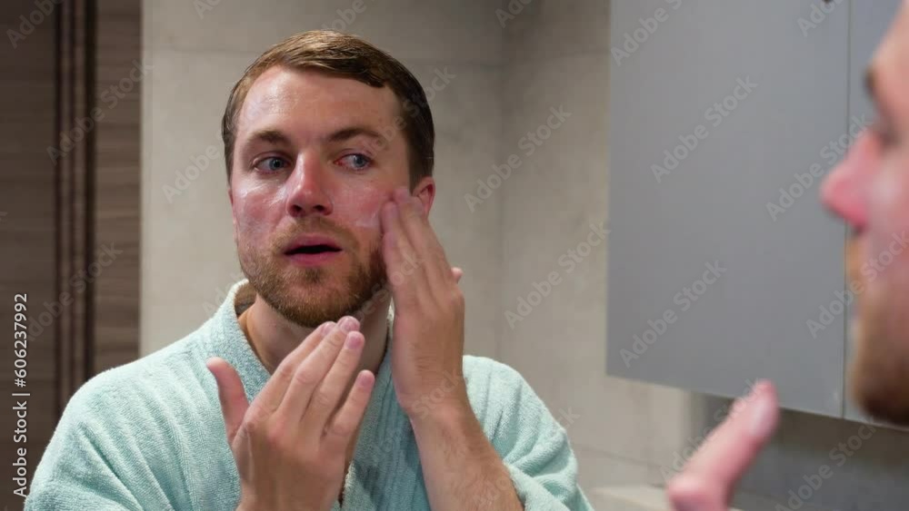 Handsome man smears face cream. A young guy looks at himself in the mirror and puts cream on his face. Men's personal care Handsome man smears face cream. A young guy looks at himself in the mirror and puts cream on his face. Men's personal care