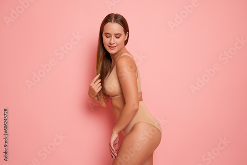 Pretty girl with long hair in toffee colour underwear posing on pink studio background in sexy pose with eyes down, comfortable underclothing concept, copy space