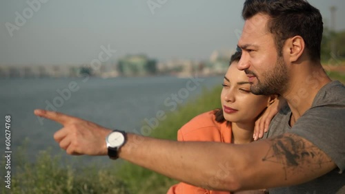 Beautiful Caucasian Couple Bonding Sitting Near River. Woman Leaning on Man's Shoulder, Man Talks And Fingerpointing Far Away. Lovers relaxing outdoors in a city park - portrait slow motion photo