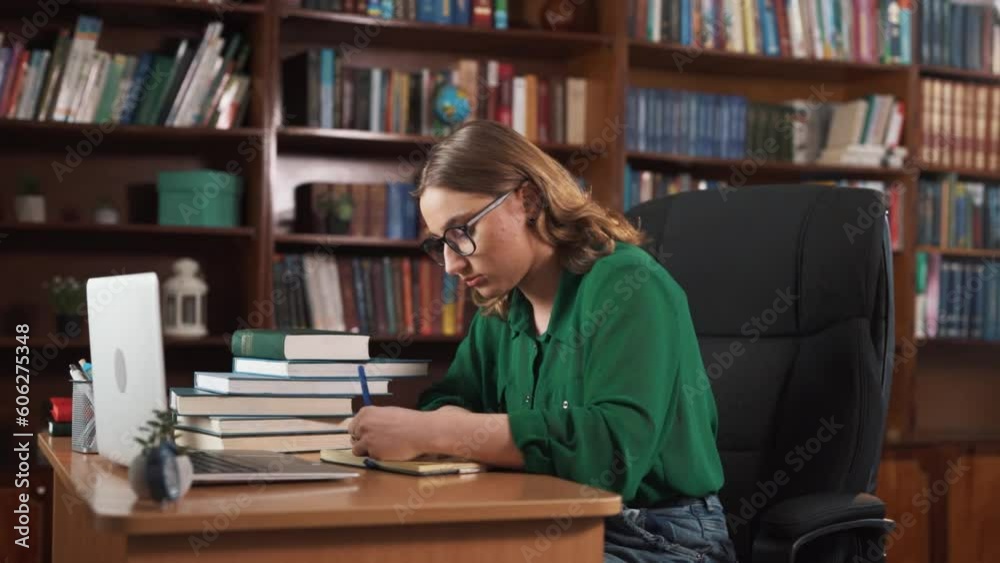 The girl is sitting at the table with several books and a laptop on it. She is writing something in her notebook and appears highly focused and thoughtful. She leans back in her chair to contemplate. The girl is sitting at the table with several books and a laptop on it. She is writing something in her notebook and appears highly focused and thoughtful. She leans back in her chair to contemplate.