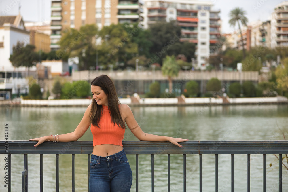 Young beautiful woman with straight brown hair, orange top and jeans, leaning on a railing looking at the ground while smiling. Concept fashion, beauty, trend, peace, relax, millennial.