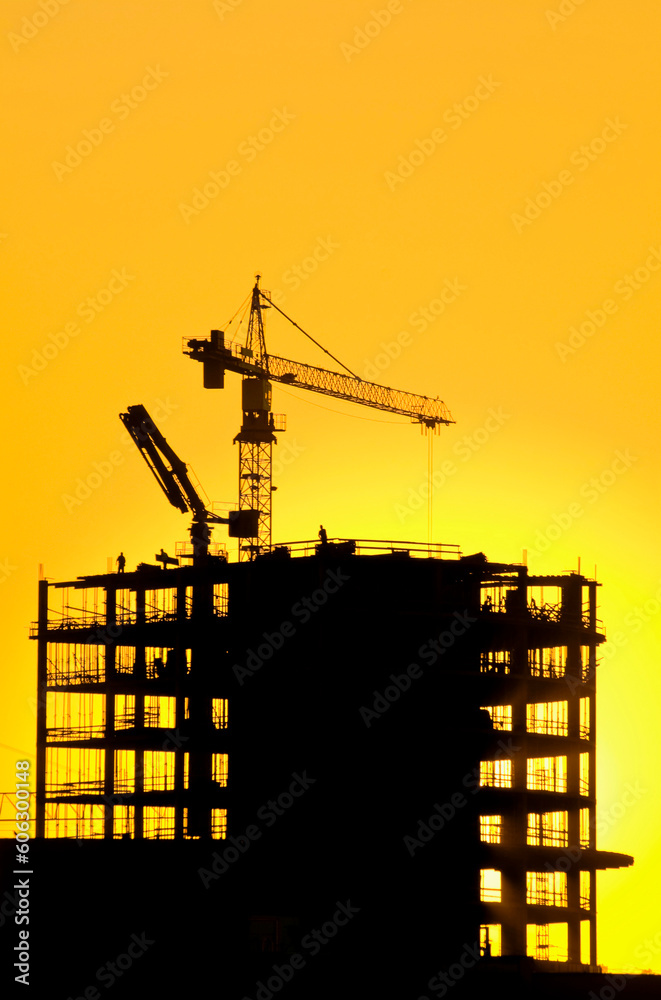 Silhouette of a skyscraper under construction process and cranes in the sunset sky. New residential buildings being constructed.