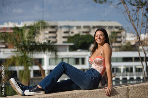 Young beautiful woman with straight brown hair, orange top and jeans, sitting on a stone wall, looking at the camera, sunbathing relaxed and happy. Concept fashion, beauty, trend, relax, millennial.