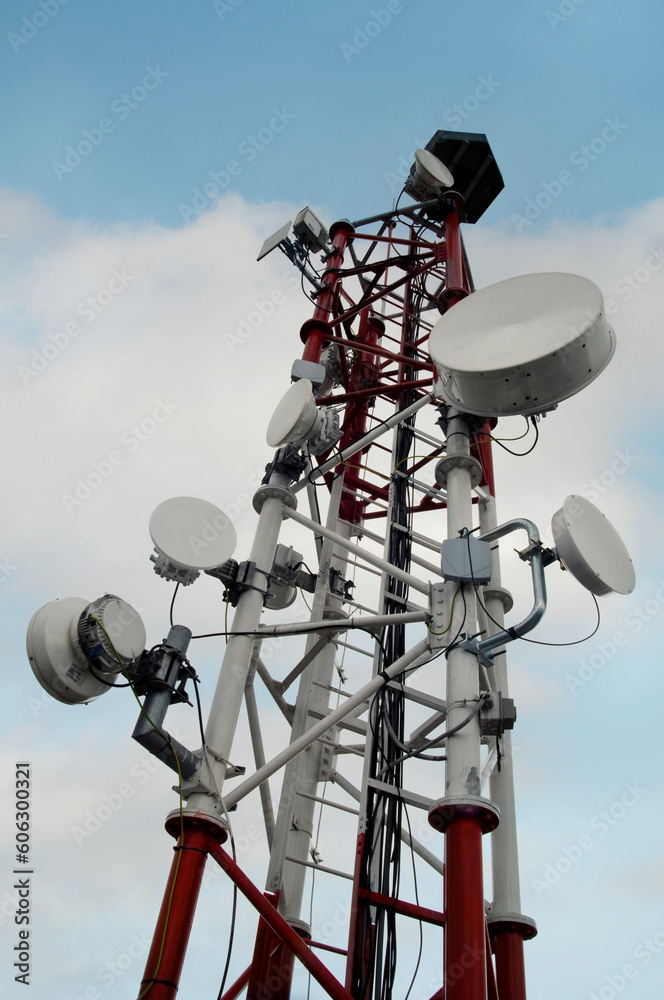 Telecommunication tower with microwave panel antennas, remote transmitter units, wireless technology, GSM, 5G, optic fibers on roof of building in blue sky with clouds.