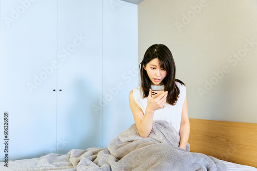 自宅の寝室で朝の寝起きにスマートフォンを見て寝坊した事にビックリし出勤に遅刻しそうな1人暮らしの女性 photo