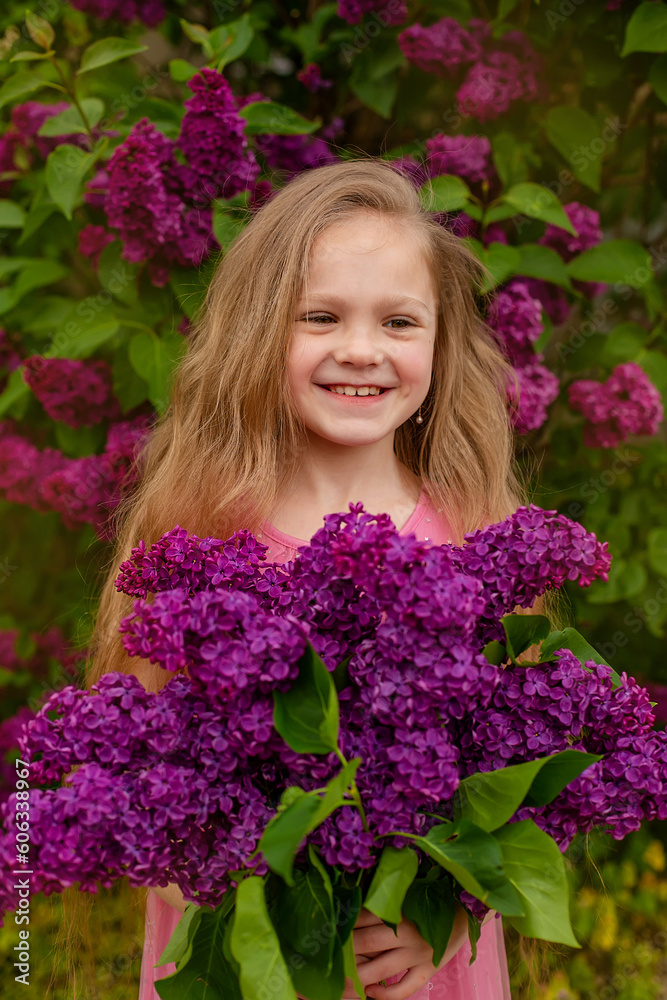 a beautiful happy smiling blonde girl with long hair in a pink dress with a purple bouquet of lilacs in her hands stands against the background of a lilac bush