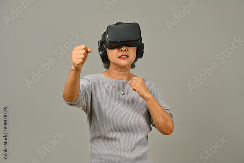 Happy senior woman in boxing stance playing action simulator game, fighting with fist in VR headset. Modern technologies, innovations