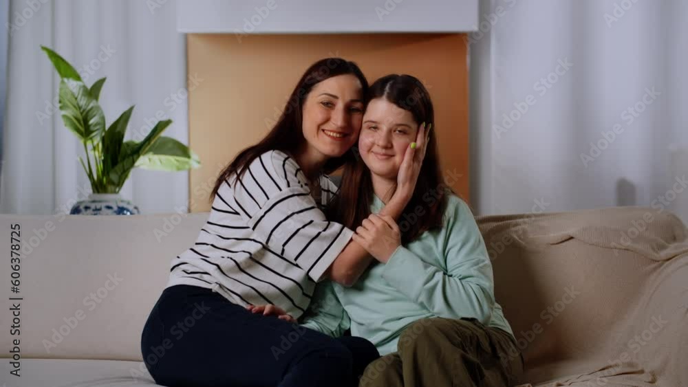 Mom and daughter sit on the couch and look at the camera with a smile. A brunette woman hugs her daughter and kisses her on the cheek. Mom and daughter sit on the couch and look at the camera with a smile. A brunette woman hugs her daughter and kisses her on the cheek.