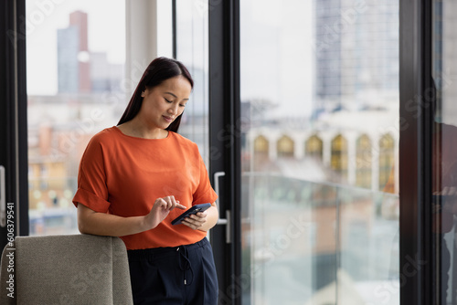 Young asian businesswoman in corporate office with view of city skyline using smartphone