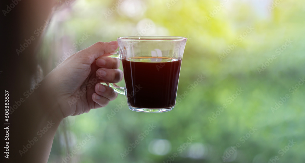 Woman hand holding and showing a glass cup of hot coffee, espresso coffee, black coffee at outdoors on green leaf background. Woman hand holding and showing a glass cup of hot coffee, espresso coffee, black coffee at outdoors on green leaf background.