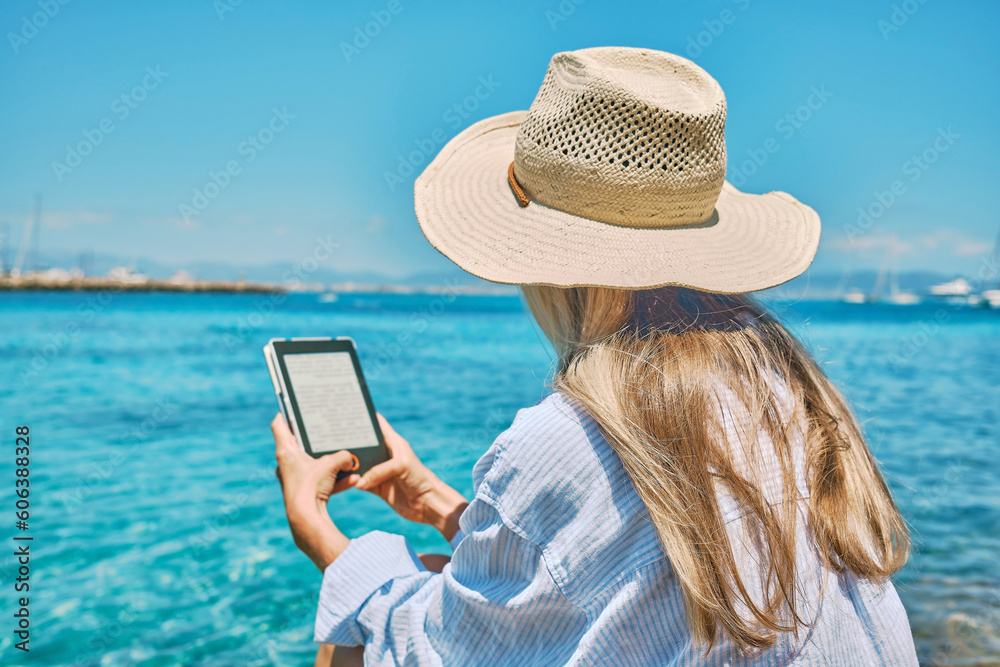 Fototapeta premium Fashion pretty woman outdoors lifestyle watching, reading on tablet ebook on the beach in summer day. Wearing wide brimmed hat, Sunbating with uv protection. Concept of beach vacation. 