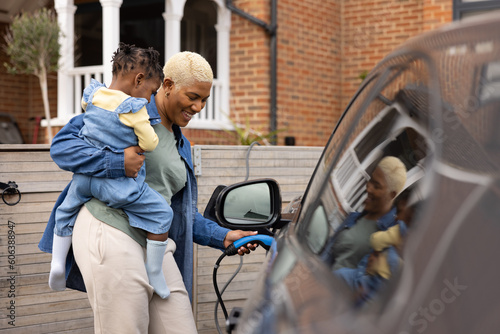 Mother charging electric car whilst holding daughter