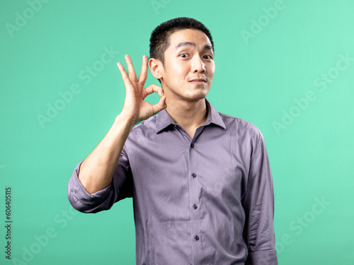 A portrait of an Asian man wearing a purple shirt, while making displaying the OK hand sign, isolated on a green background.