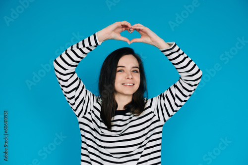 Joyful Woman in Striped Sweater Spreading Love - Smiling Girl with Raised Arms Creating Heart Shape, Radiating Happiness and Positivity - Isolated Blue Background - Perfect for Positive Vibes Concepts © Yauhen