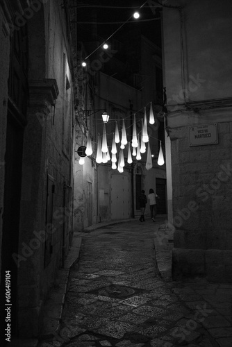 Grayscale vertical shot of a narrow stone ally at night with hanging lights
