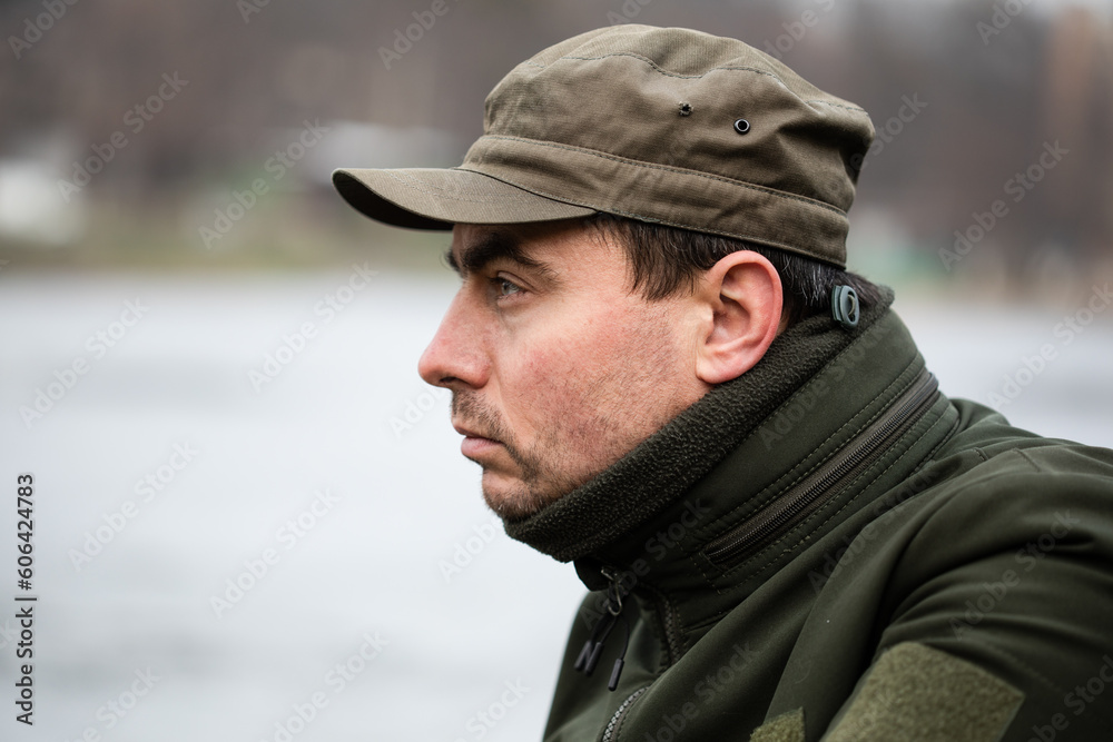 Portrait of serious military man in olive uniform and cap against lake in rural area on cold winter day