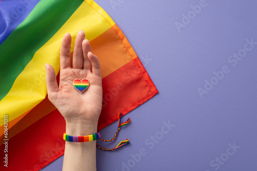A first person top view of symbolic rainbow-colored bracelet adorns a girl's hand holding a heart-shaped badge over flag on a lilac background with space for text, recognizing LGBTQ History Month photo