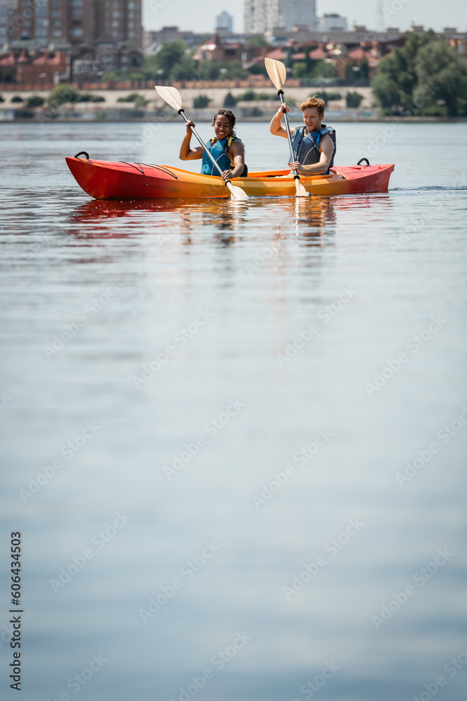 positive and active multiracial couple in life vests holding paddles while sailing in sportive kayak on calm water surface during summer recreation weekend on city lake positive and active multiracial couple in life vests holding paddles while sailing in sportive kayak on calm water surface during summer recreation weekend on city lake
