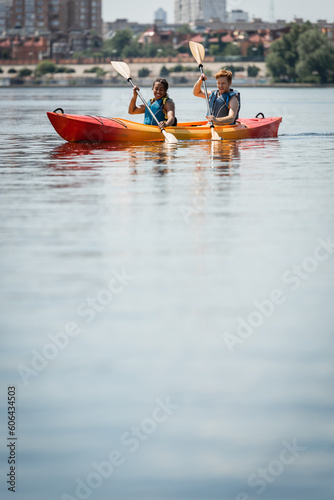 positive and active multiracial couple in life vests holding paddles while sailing in sportive kayak on calm water surface during summer recreation weekend on city lake