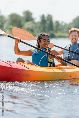 carefree and charming african american woman in life vest holding paddle and sailing in sportive kayak near young redhead man smiling on blurred background