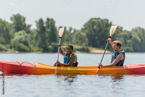joyful and active interracial couple in life vests paddling in sportive kayak while spending time on lake with blurred green shore during summer vacation