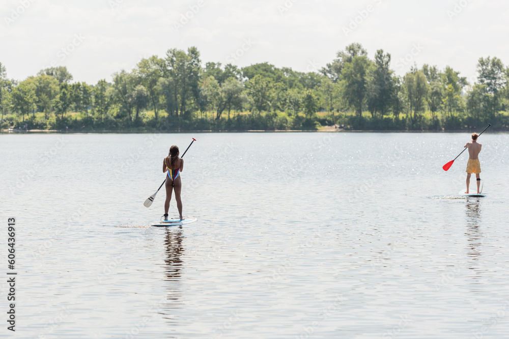 back view of sportive man and african american woman in colorful swimwear sailing on sup boards with paddles on lake with green picturesque shore, outdoor activity, summer vibes
