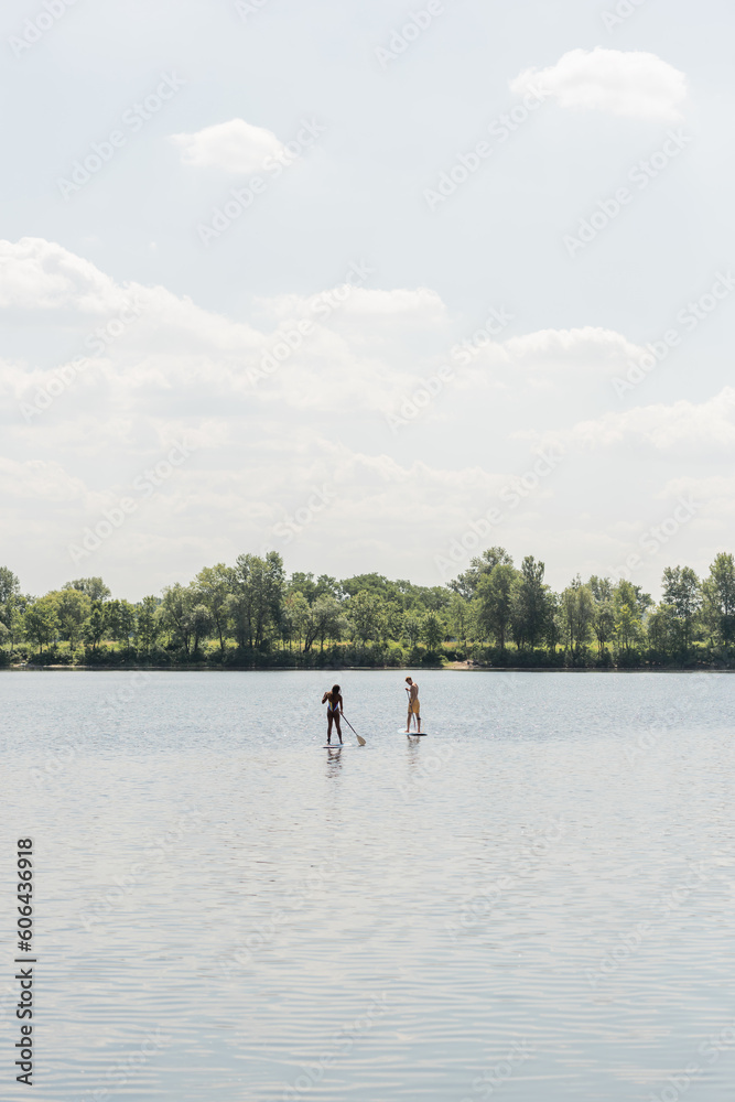 view from afar of sportive interracial couple sailing on sup boards while spending weekend on lake with green picturesque shore under cloudy sky, outdoor activity, summer vibes