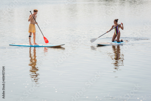 sportive african american woman in colorful swimsuit and young redhead man in swim shorts spending summer weekend while sailing on sup boards together © LIGHTFIELD STUDIOS