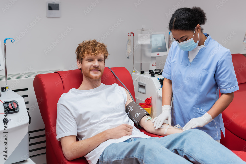 Obraz premium redhead volunteer sitting on medical chair near automated equipment while multiracial nurse in medical mask fixing transfusion set in blood donation center
