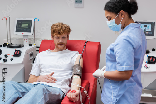 redhead volunteer with transfusion set squeezing rubber ball while sitting on medical chair near automated equipment and multiracial nurse in medical mask in blood donation center
