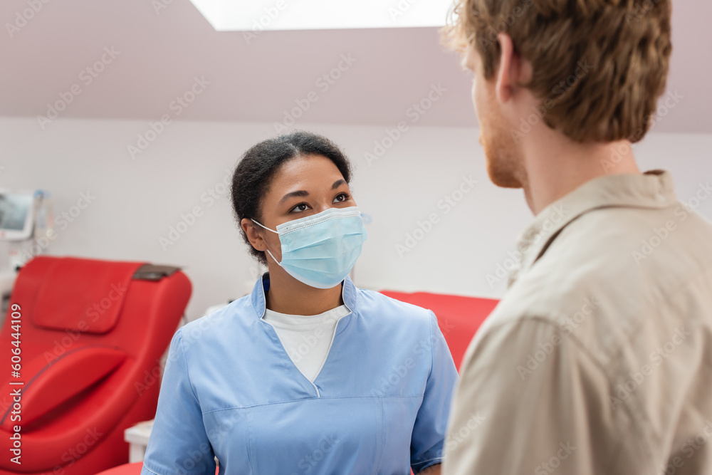 multiracial healthcare worker in blue uniform and medical mask talking to young redhead volunteer on blurred foreground near medical chairs in blood donation center