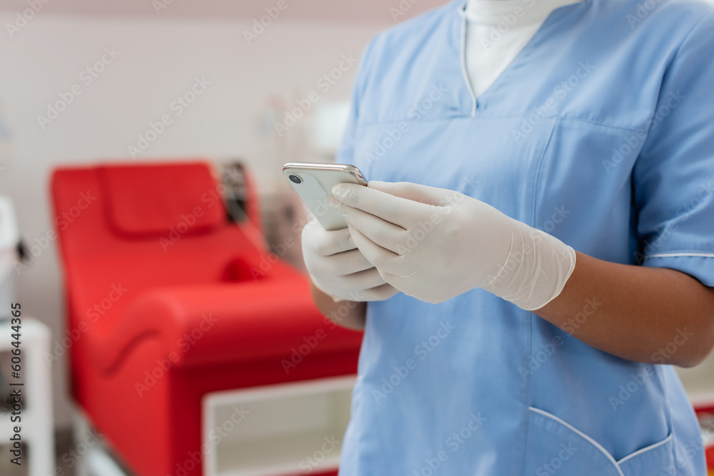 partial view of multiracial nurse in blue uniform and sterile latex gloves messaging on mobile phone near blurred medical chair in blood transfusion center partial view of multiracial nurse in blue uniform and sterile latex gloves messaging on mobile phone near blurred medical chair in blood transfusion center