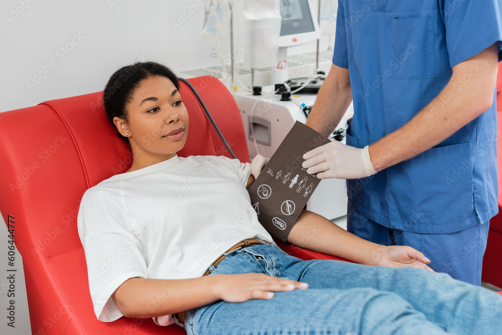 healthcare worker in blue uniform and latex gloves putting blood pressure cuff on arm of multiracial woman sitting on comfortable medical chair near automated transfusion machine in hospital healthcare worker in blue uniform and latex gloves putting blood pressure cuff on arm of multiracial woman sitting on comfortable medical chair near automated transfusion machine in hospital