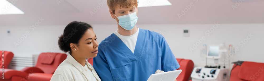 multiracial woman and redhead practitioner in blue uniform and medical mask looking at digital tablet near medical chairs and blood transfusion machine in clinic, banner multiracial woman and redhead practitioner in blue uniform and medical mask looking at digital tablet near medical chairs and blood transfusion machine in clinic, banner