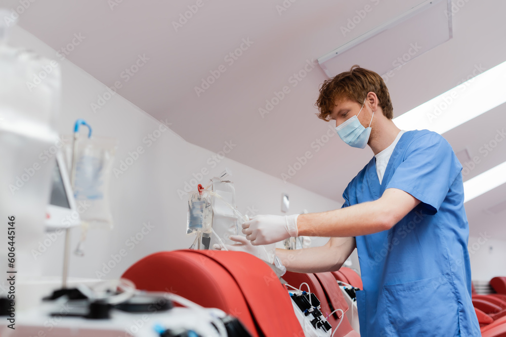 redhead healthcare worker in medical mask and latex gloves assembling transfusion set near automated equipment and medical chairs in blood donation center redhead healthcare worker in medical mask and latex gloves assembling transfusion set near automated equipment and medical chairs in blood donation center