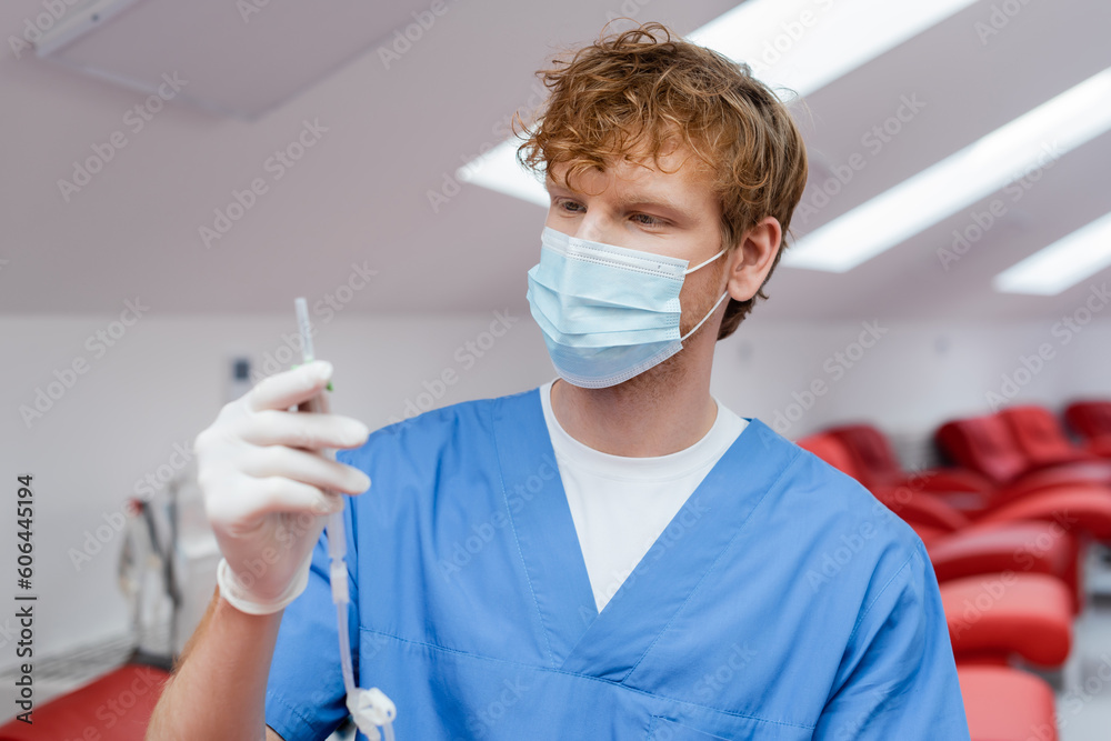 young and redhead doctor in blue uniform, medical mask and latex glove holding blood transfusion set near ergonomic medical chairs on blurred background in hospital young and redhead doctor in blue uniform, medical mask and latex glove holding blood transfusion set near ergonomic medical chairs on blurred background in hospital